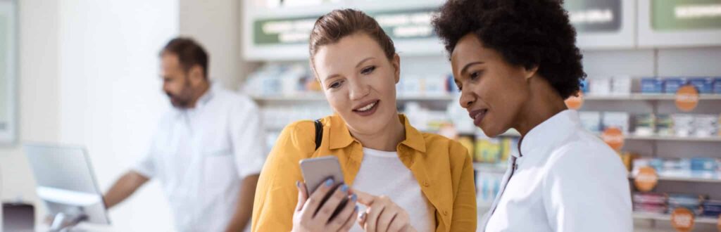 Two smiling women looking at a phone together in a pharmacy, with a pharmacist working in the background.