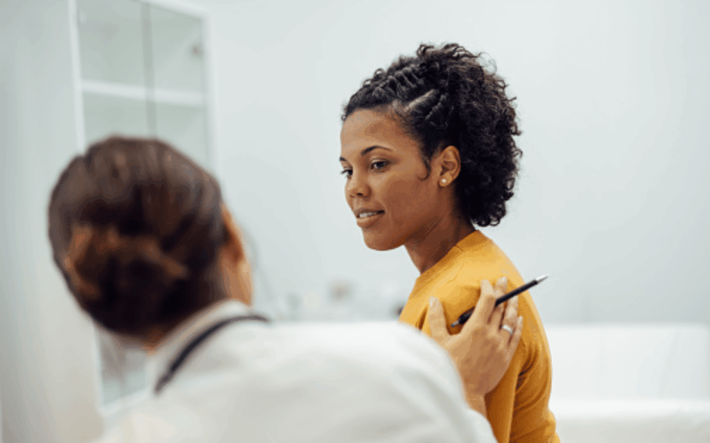Woman in a yellow shirt smiling as a doctor places a hand on her shoulder in an exam room.